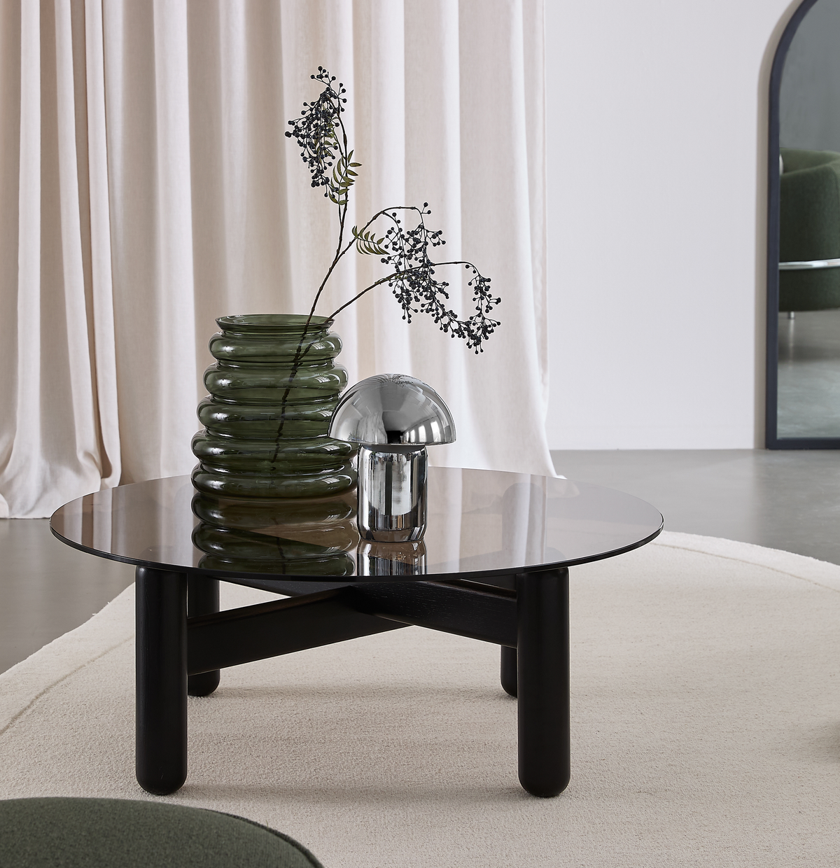 Rundt sort sofabord med tonet glasplade, grøn rillet glasvase med gren og lille krom svampelampe på et beige tæppe foran lyse gardiner i en minimalistisk stue.