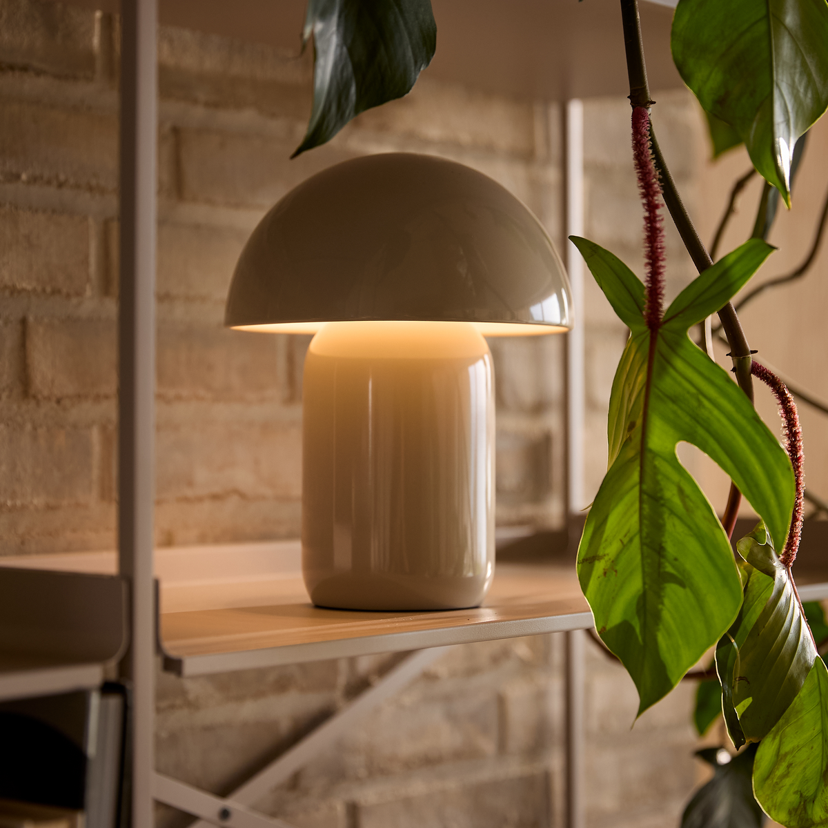 Svampeformet beige bordlampe lyser varmt på en hylde ved siden af en grøn klatreplante mod en murstensvæg.