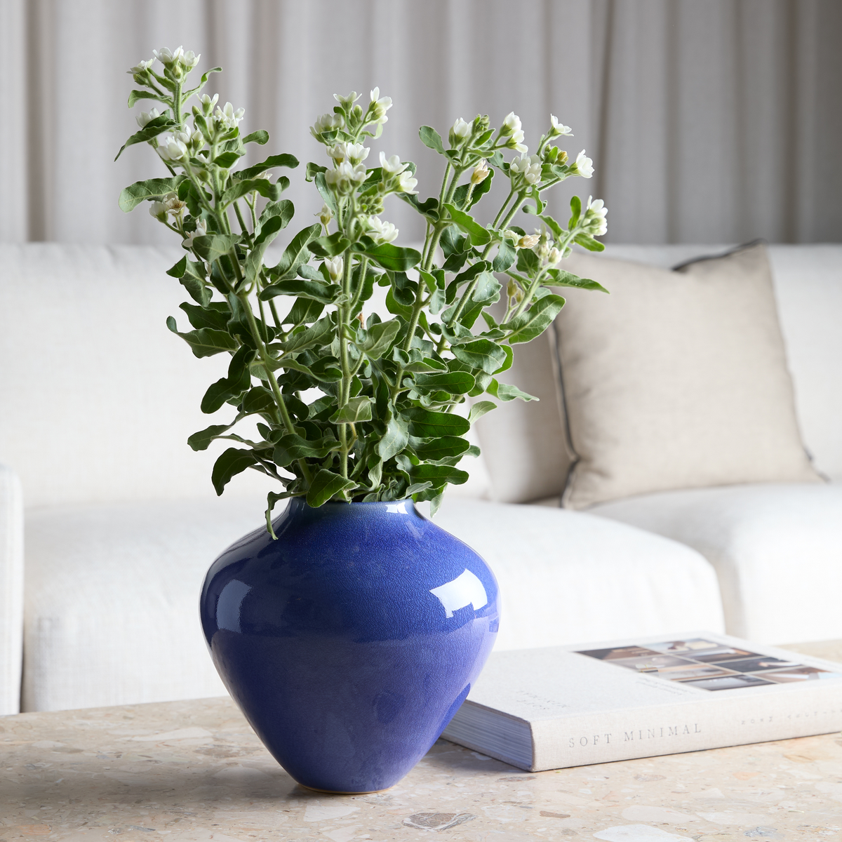 Blå vase med grønne blade og hvide knopper på et stenbord ved siden af bogen Soft Minimal i en lys stue.