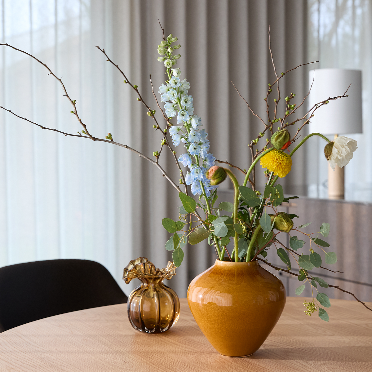 Buket i sennepsgul keramikvase på lyst træbord med blå, gule og hvide blomster og grene; lille ravfarvet glasdekoration ved siden af, bløde gardiner og bordlampe i baggrunden.