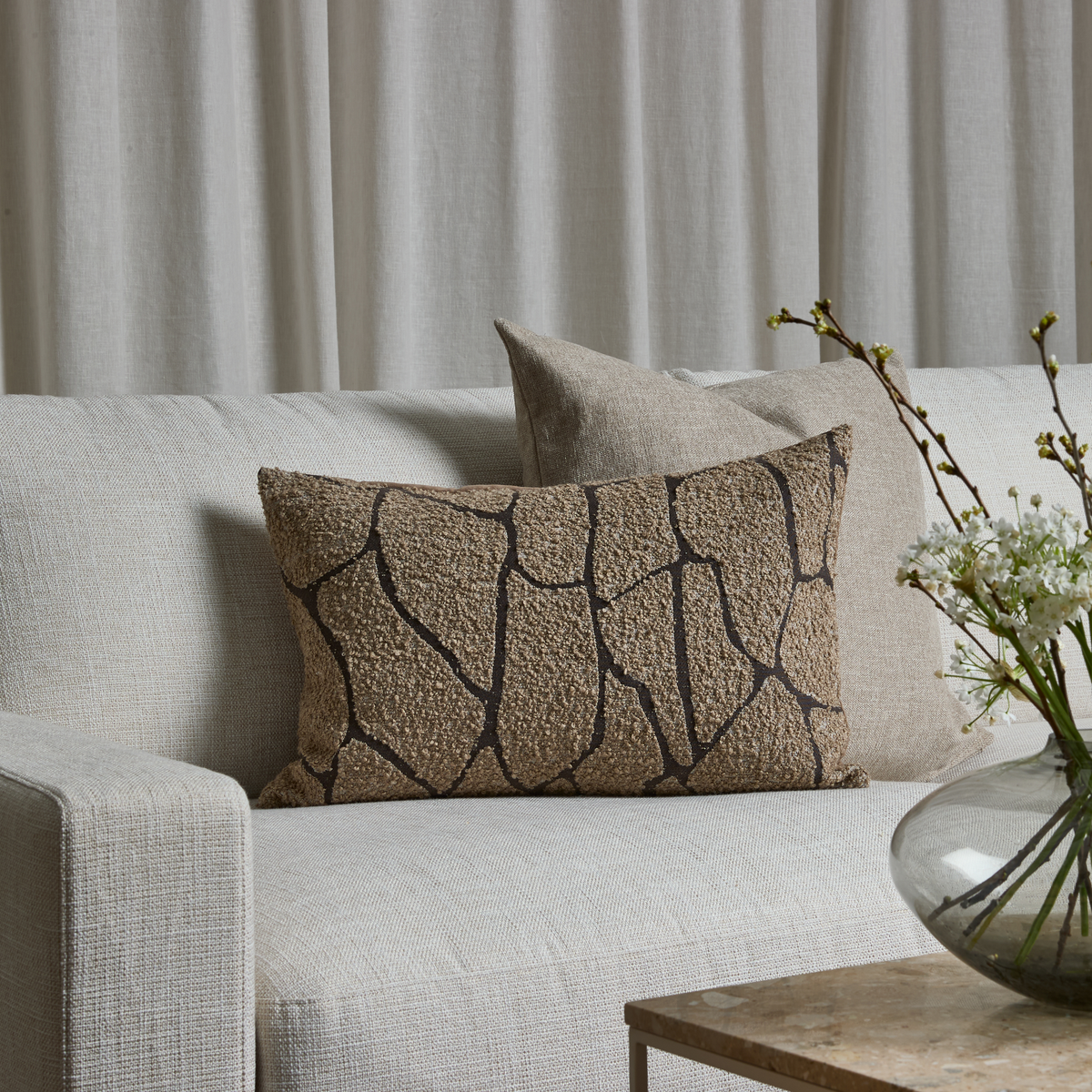 Beige sofa med to puder, en brunmønstret aflang pude foran en ensfarvet beige; lyse gardiner bagved og en glasvase med hvide blomster på et bord.