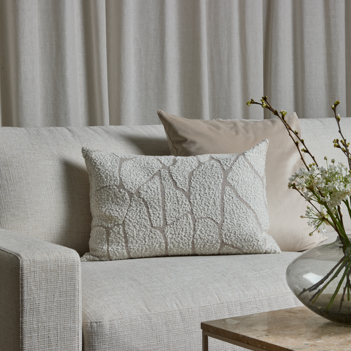 Nærbillede af en lys beige sofa med en hvid tekstureret mønstret aflang pude og en glat beige pude foran lyse gardiner samt en glasvase med hvide blomster på et stenbord.