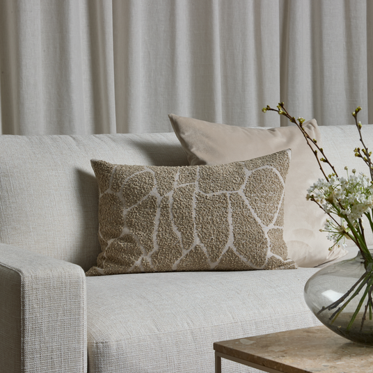 Beige, tekstureret pude på lys sofa med neutrale puder, linnedgardiner og glasvase med grene og hvide blomster på et stenbord.