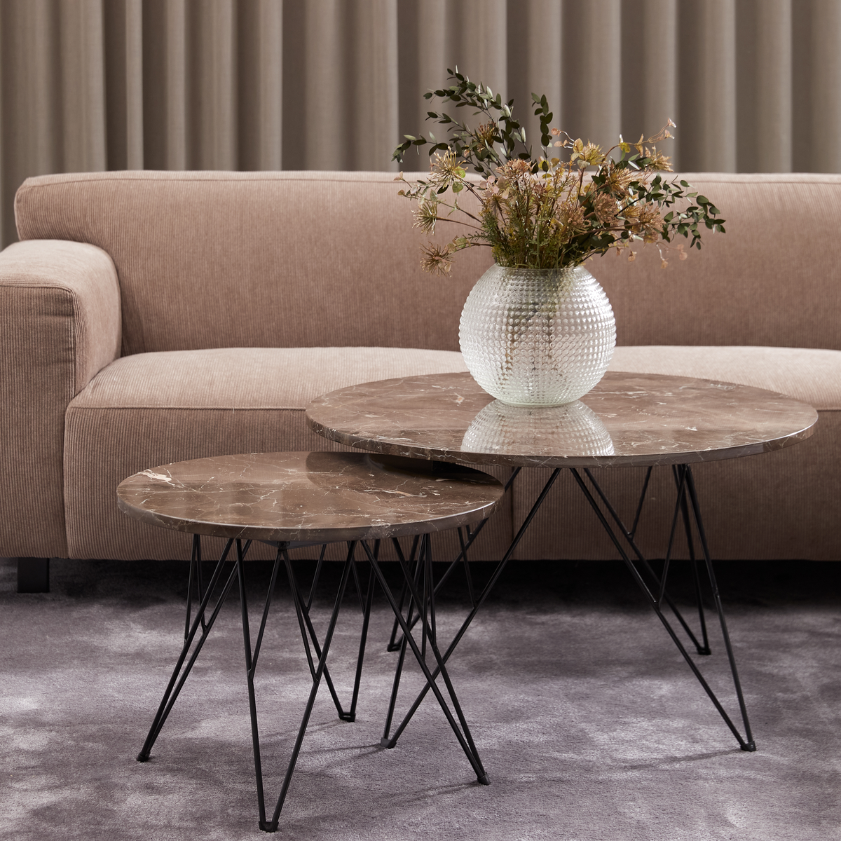 Beige sofa med to runde sofaborde i brun marmor med sort stel; glasvase med tørrede blomster på det større bord, gråt tæppe og beige gardiner.