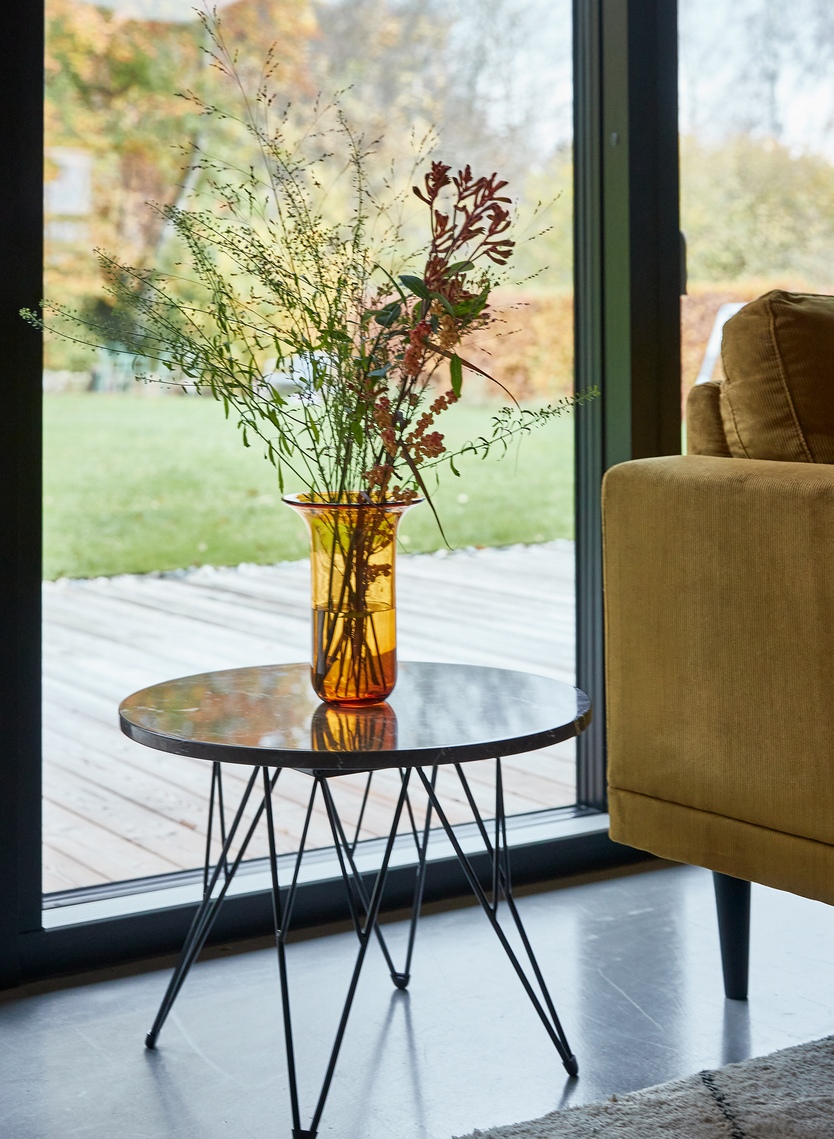Rundt sidebord i metal med ravfarvet vase med vilde grene ved siden af en sennepsgul sofa ved store vinduer med udsigt til træterrasse og græsplæne.