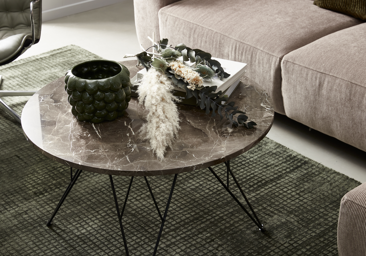 Rundt sofabord i brun marmor med sort stel, grøn vase, bøger og tørrede planter på grøn tæppe ved siden af beige sofa.