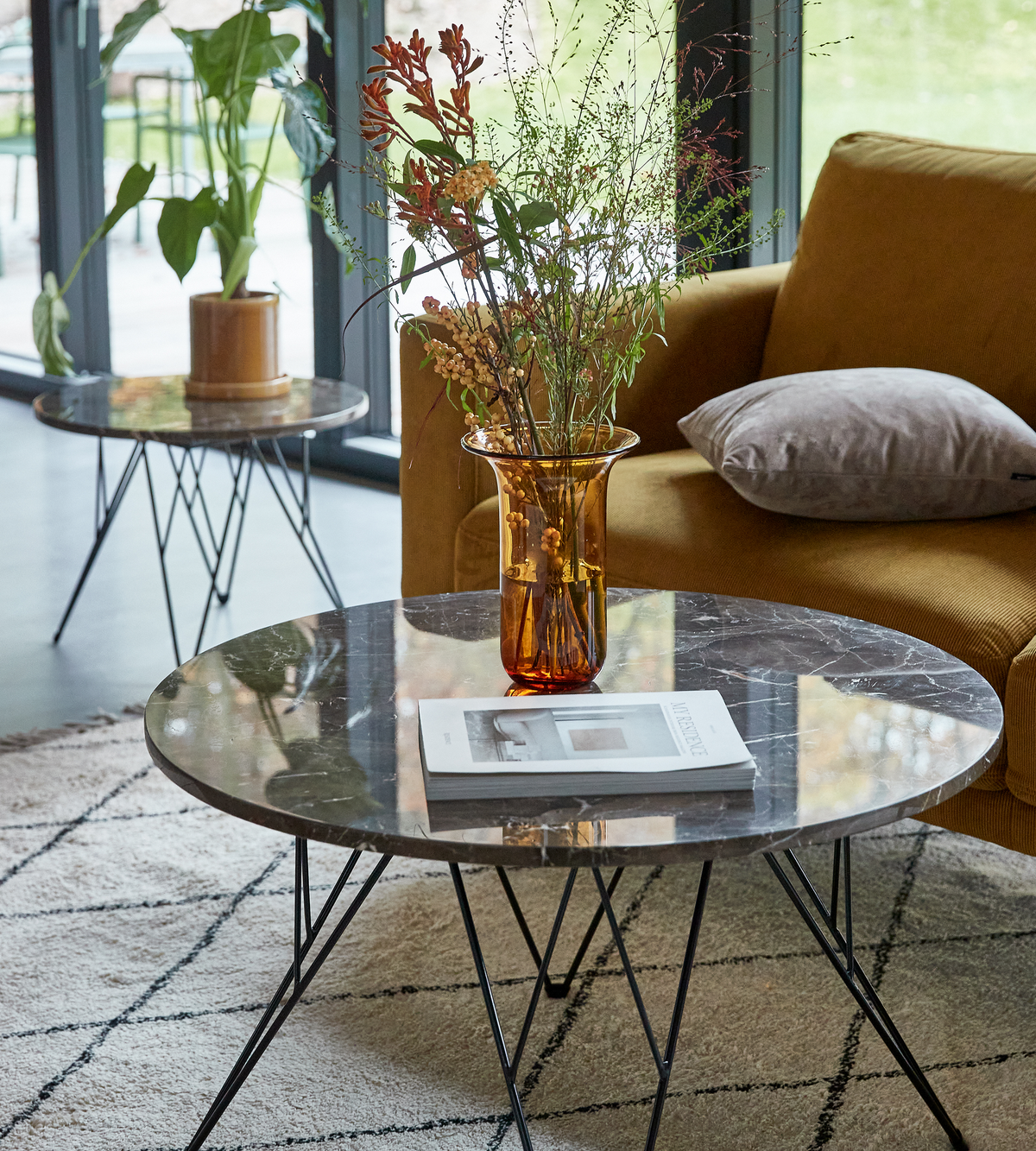 Rundt sofabord i mørk marmor med ravfarvet vase med vilde grene, et magasin på bordet og en sennepsgul sofa med grå pude i en lys stue med store vinduer.