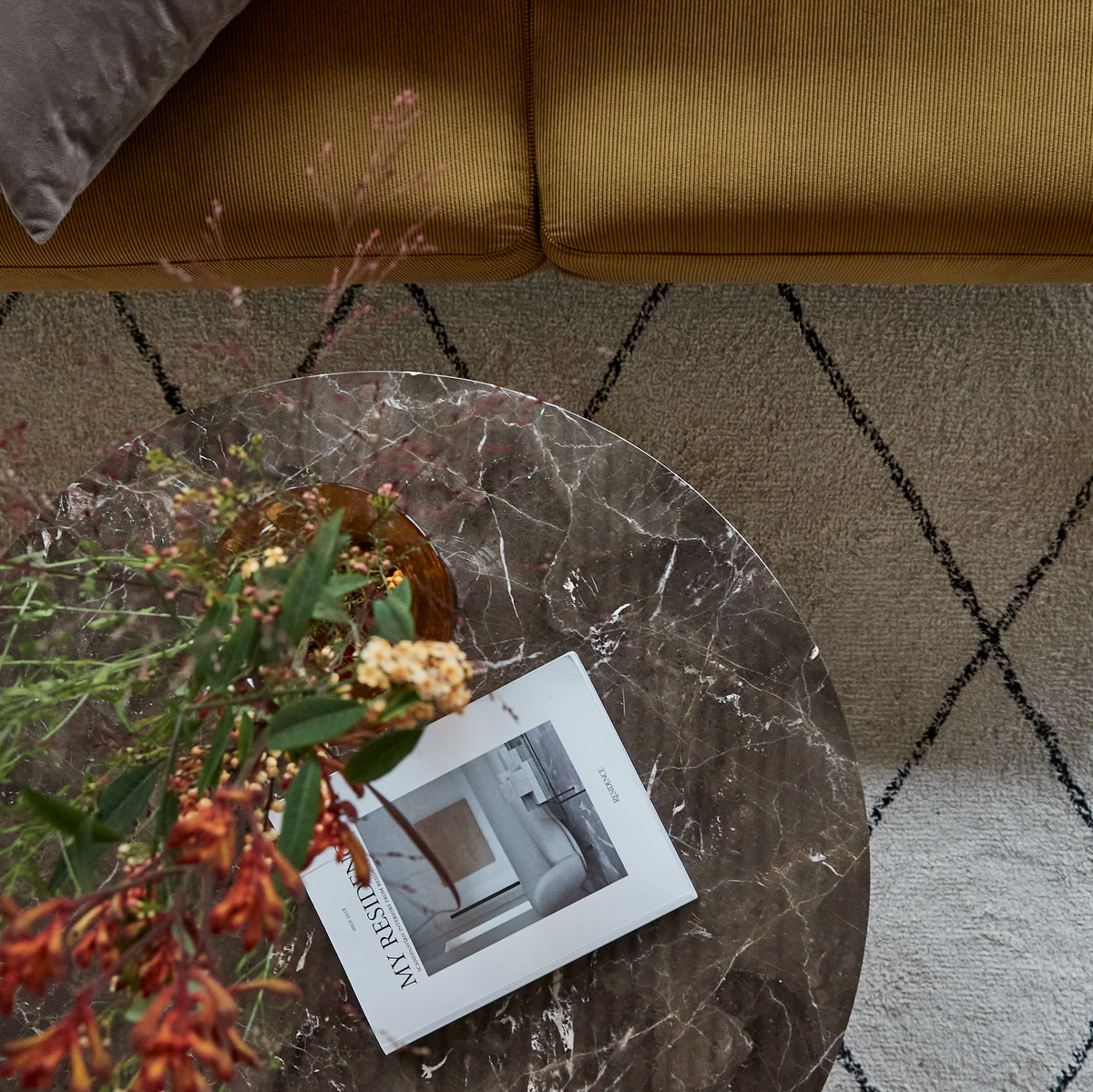 Rundt marmorbord med vase med blomster og magasinet ”MY RESIDENCE” på et beige mønstret tæppe ved siden af en sennepsgul sofa.