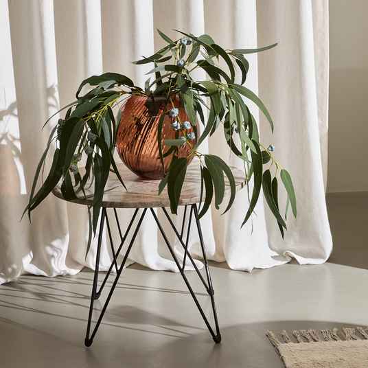 Eukalyptus i ravfarvet glasvase på rundt sidebord med hårnålsben foran lyse gardiner i sollys.