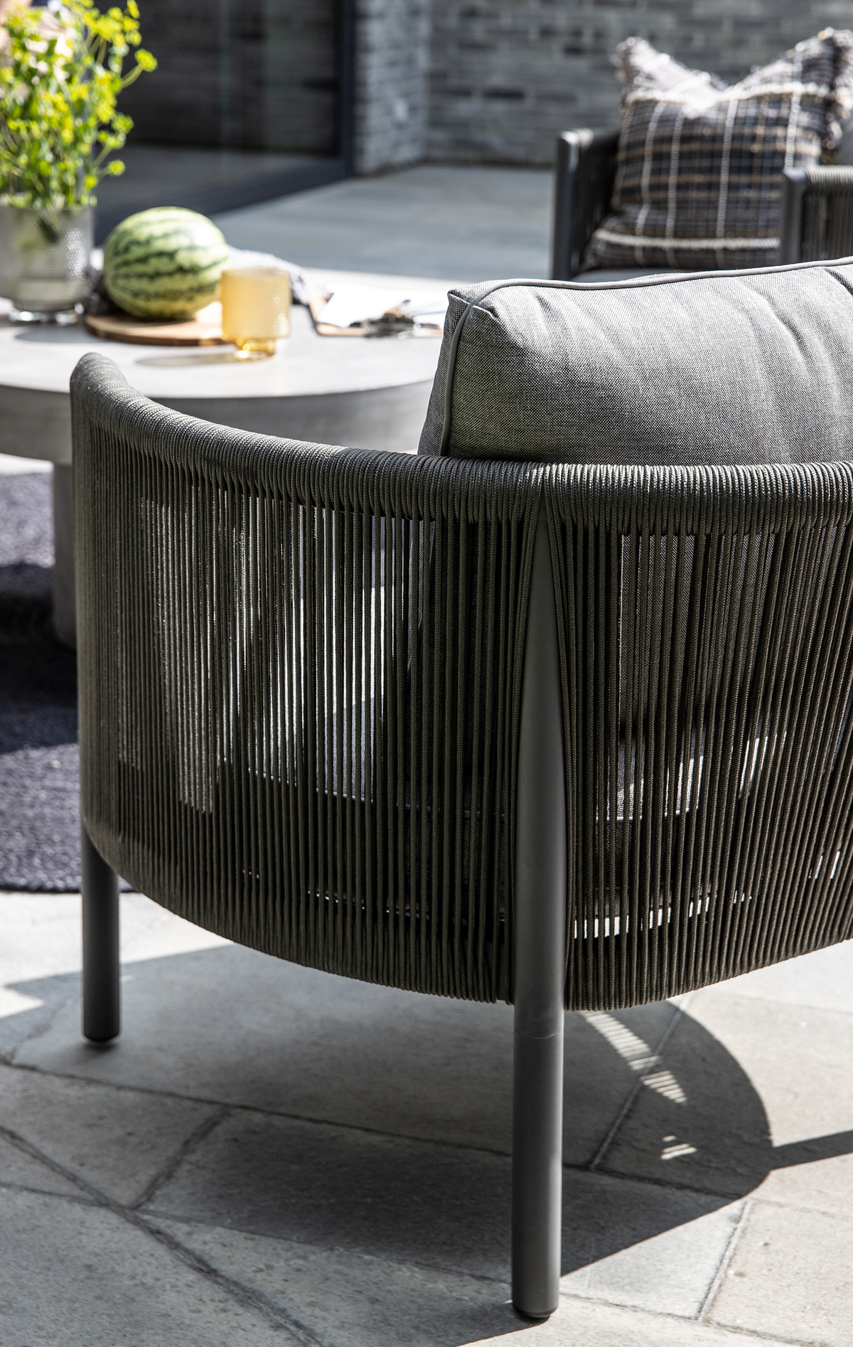 Nærbillede af udendørs lænestol med mørkt snore-design og grå pude på stenbelagt terrasse, med rundt betonbord, glas og vandmelon i sollys.