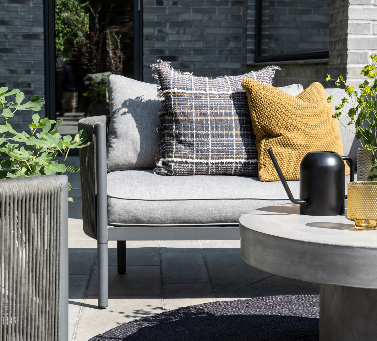 Udendørs lounge med grå lænestol, gule og ternede puder, rundt betonbord med sort vandkande og gult glas, omgivet af grønne planter i dagslys.