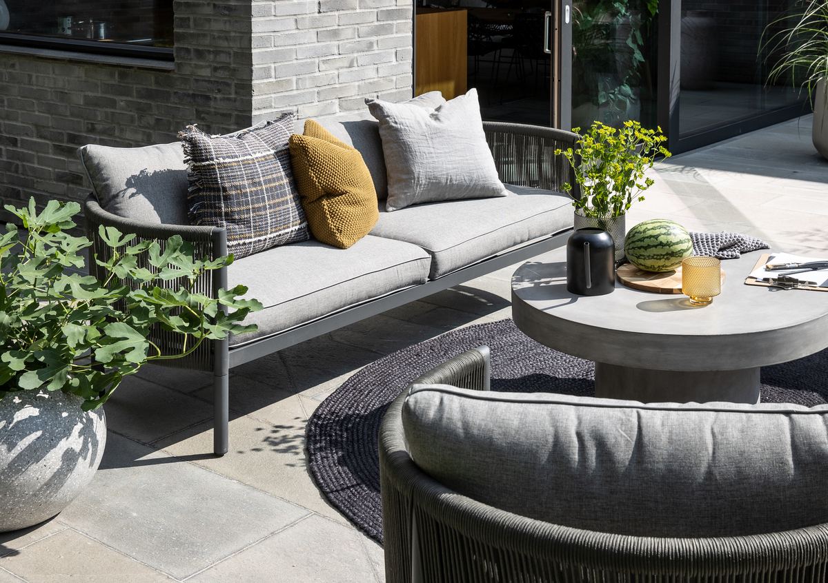 Grå udendørs sofa med puder på en terrasse, rundt betonbord med vase med grønne kviste, sort kande, glas og en hel vandmelon på bakke; krukkeplanter og mørkt rundt udendørstæppe i dagslys.