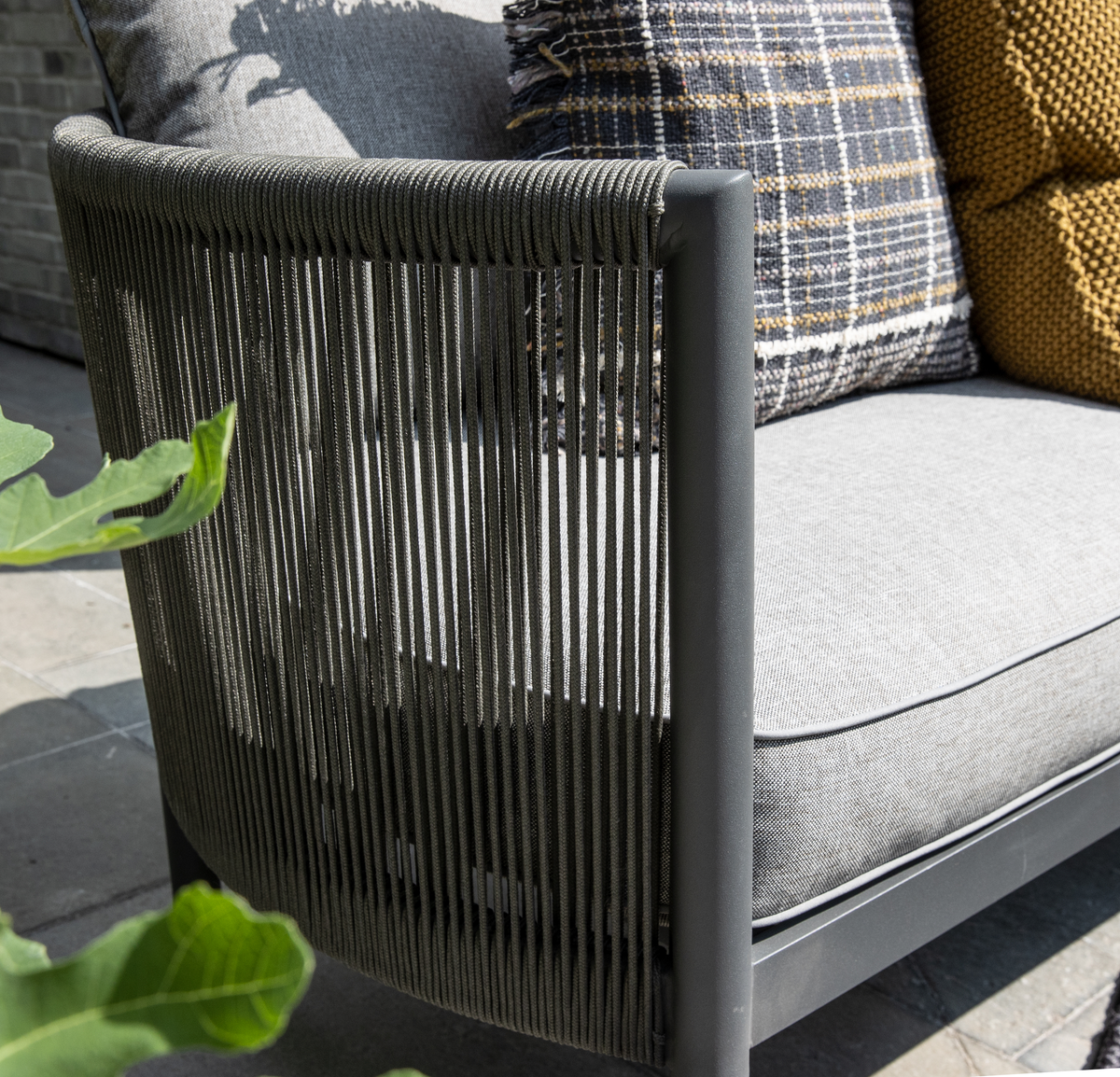 Nærbillede af en moderne udendørs sofa med grå rebdetaljer på armlænet, grå hynder samt en ternet og sennepsgul pude på en solbeskinnet terrasse.