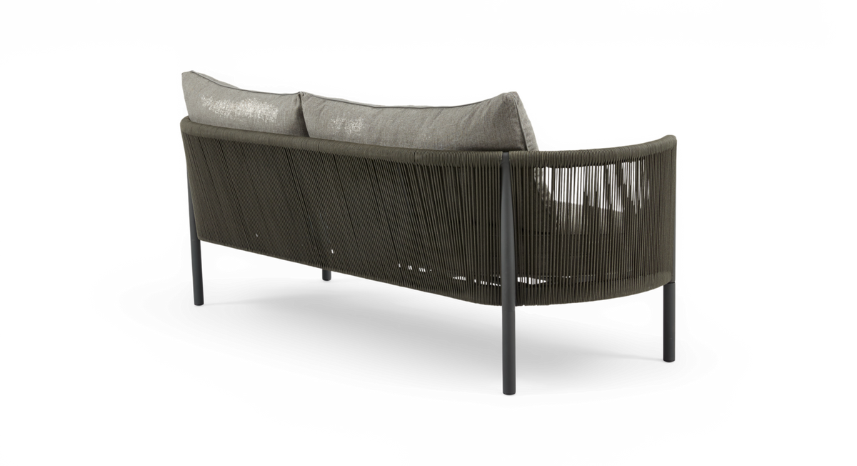 Udendørs sofa med afrundet ryg i olivfarvet rebflet og grå puder, set skråt bagfra.
