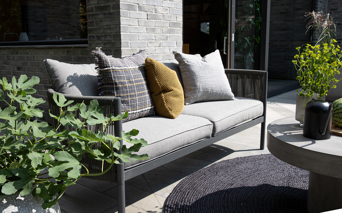 Udendørs grå sofa med puder i grå, ternet og sennepsgul, omgivet af potteplanter ved et rundt betonbord med sort kande.