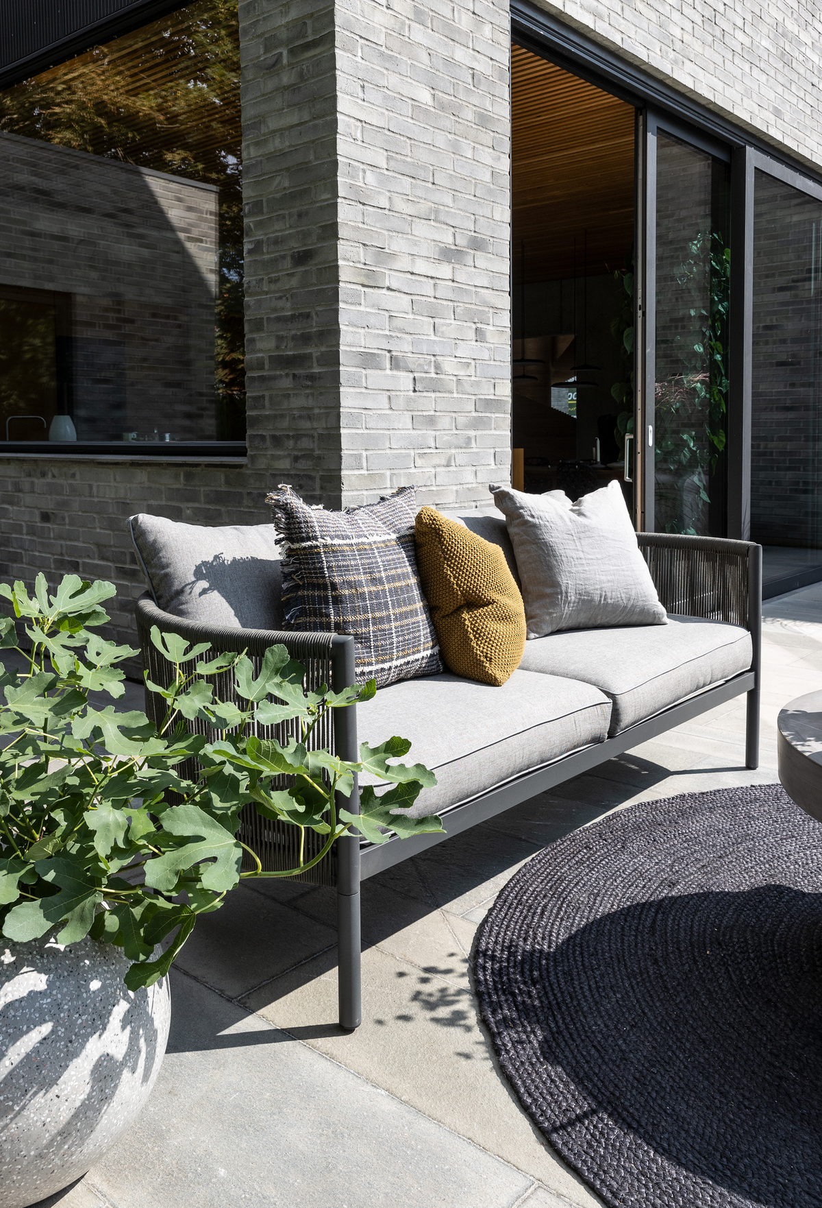 Moderne udendørs sofa med grå hynder og puder i grå, ternet og sennepsgul på en flisebelagt terrasse ved en murstensfacade, med stor potteplante og rund mørk udendørsmåtte i sollys.
