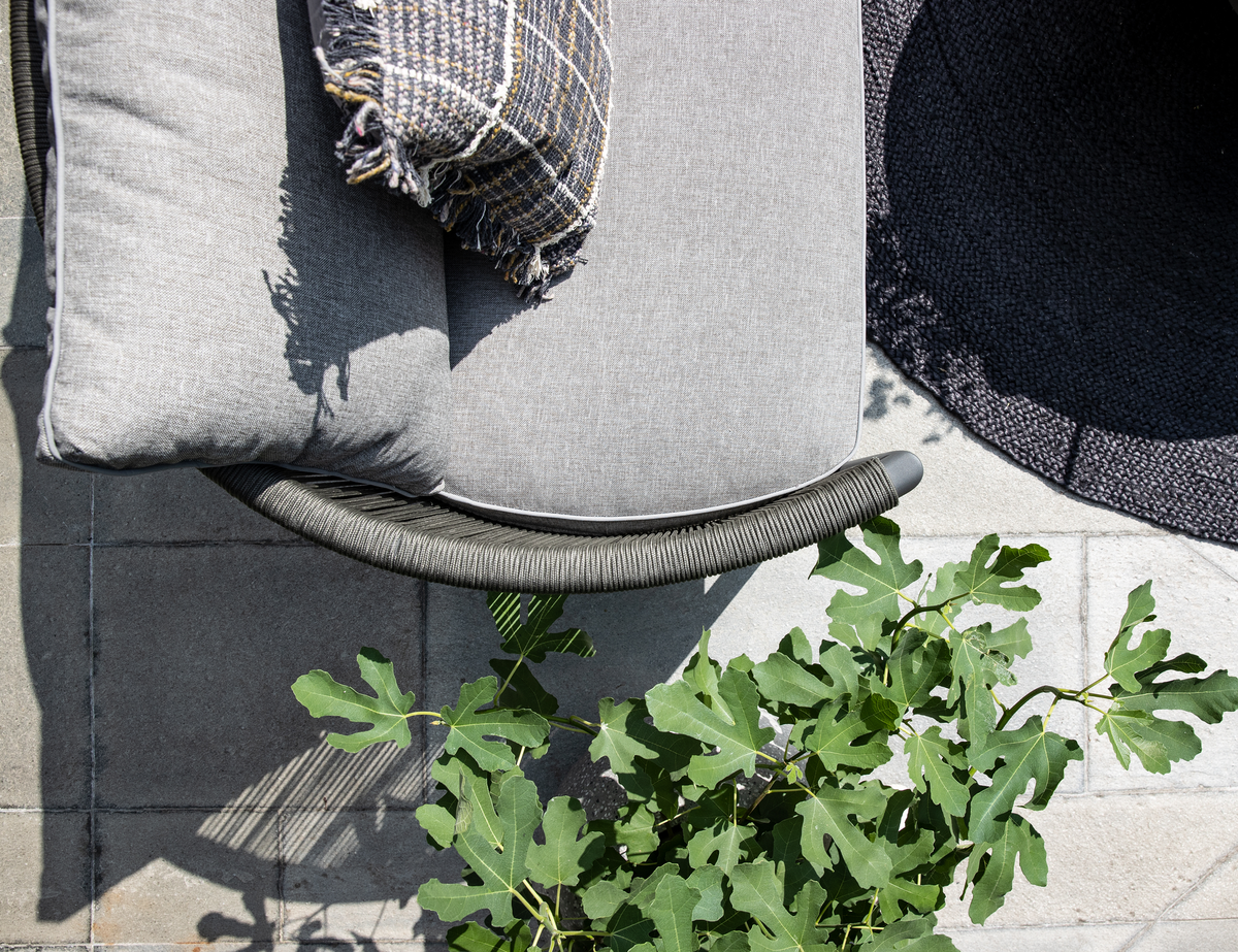 Nærbillede af grå udendørs loungestol med pude og ternet plaid, grønne fikonblade og mørk rund tæppe på flisebelagt terrasse i sollys.