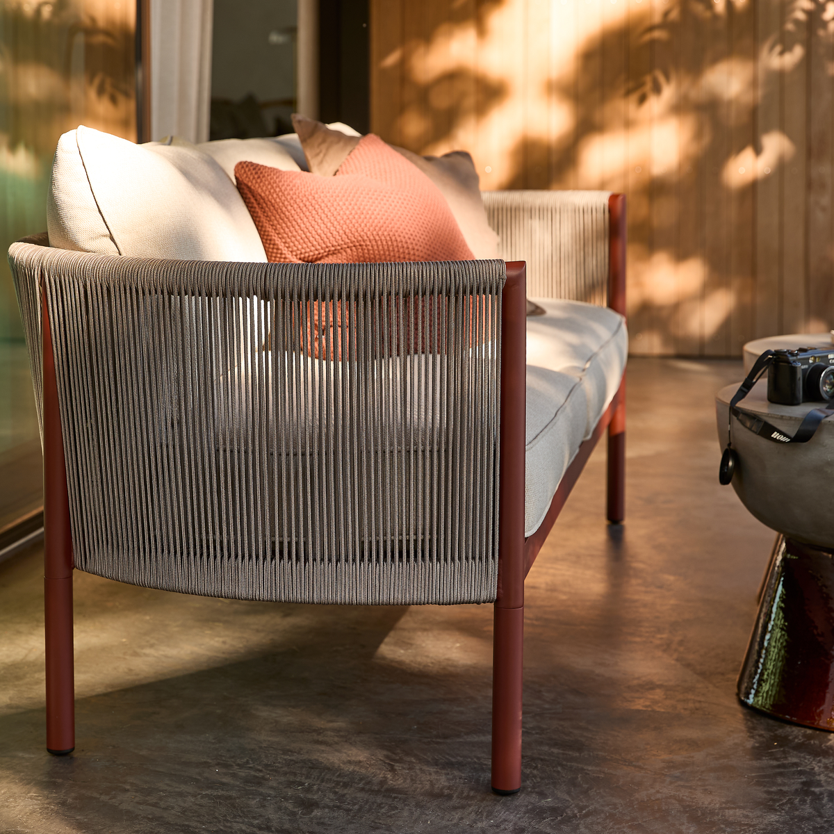 Moderne udendørs sofa med grå snor-ryg, rustrøde ben og beige hynder med terrakottafarvet pude på terrasse i sollys; kamera på sidebord.