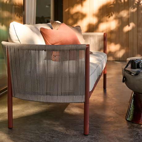 Moderne udendørs sofa med grå snor-ryg, rustrøde ben og beige hynder med terrakottafarvet pude på terrasse i sollys; kamera på sidebord.