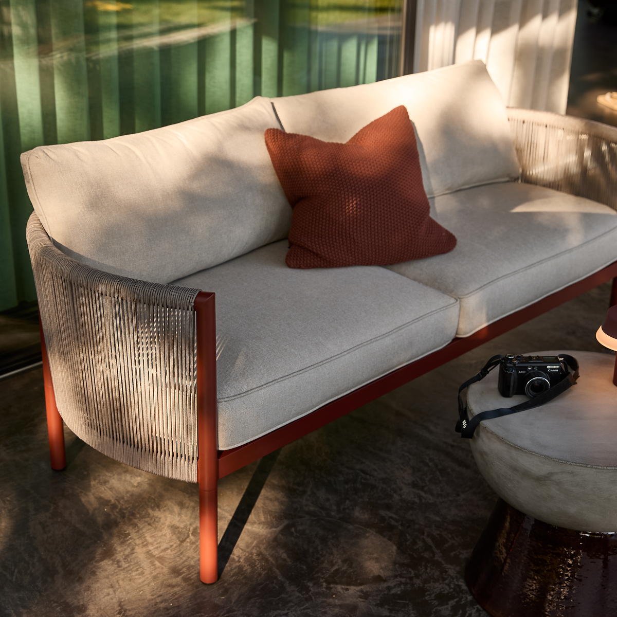 Beige udendørssofa med rust­rød ramme og snørede sider, med en terrakottafarvet pude; ved siden af står en lille puf/bord med en lille lampe og et kamera i varm aftensol.
