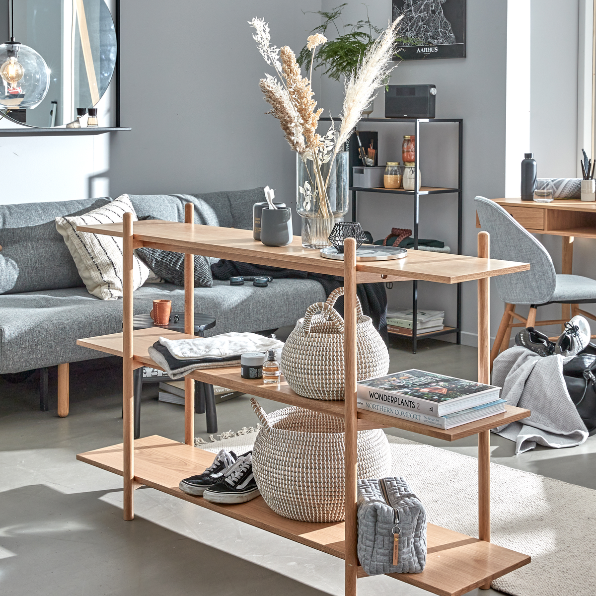 Lyst skandinavisk stue med en åben hylde i egetræ stylet med kurve, sneakers og en vase med tørrede strå; bøgerne “WONDERPLANTS 2” og “NORTHERN COMFORT” ligger på midterhylden; grå sofa og skrivebord i baggrunden.