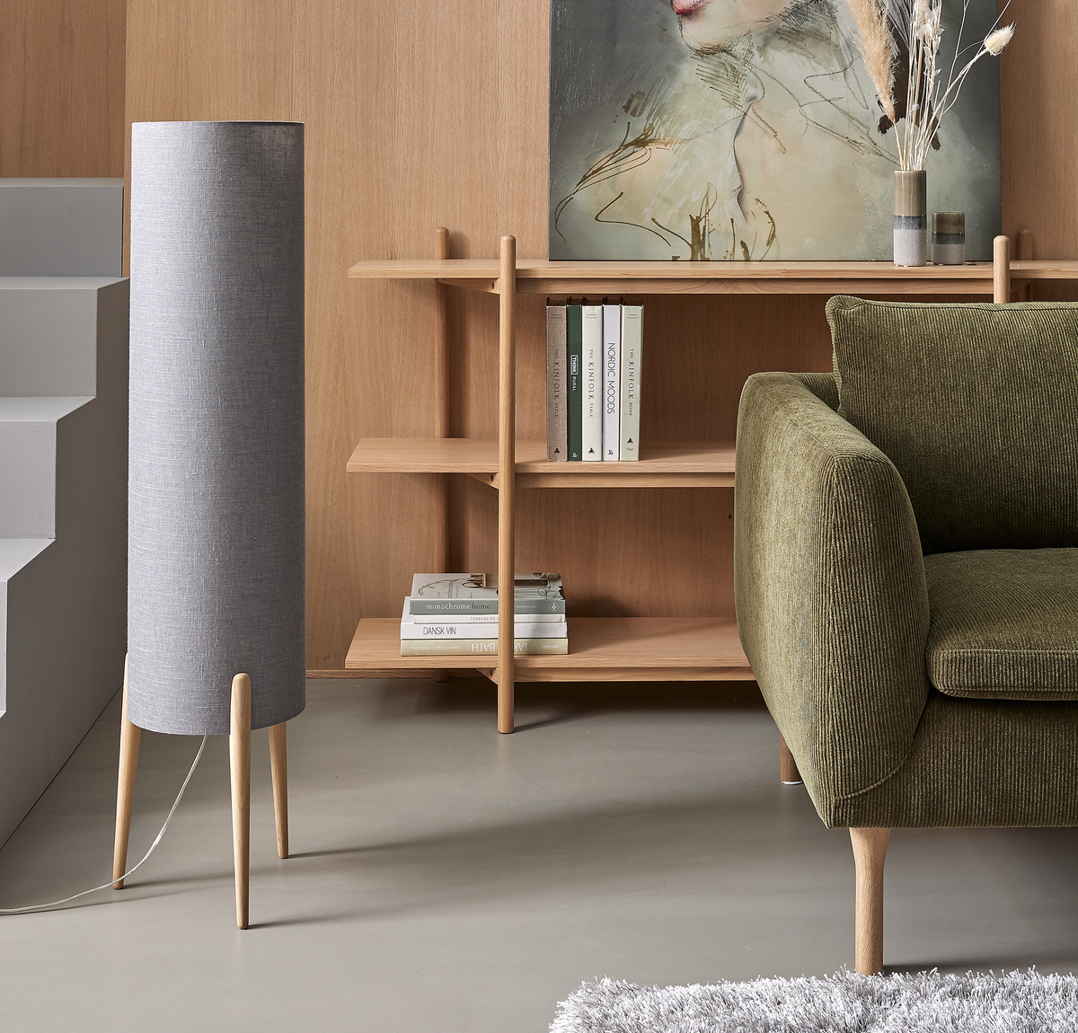 Lyst skandinavisk stue med grå cylindrisk gulvlampe, grøn sofa, egetræshylde med bøger som Nordic Moods og Dansk, vase med tørrede strå og portrætmaleri.