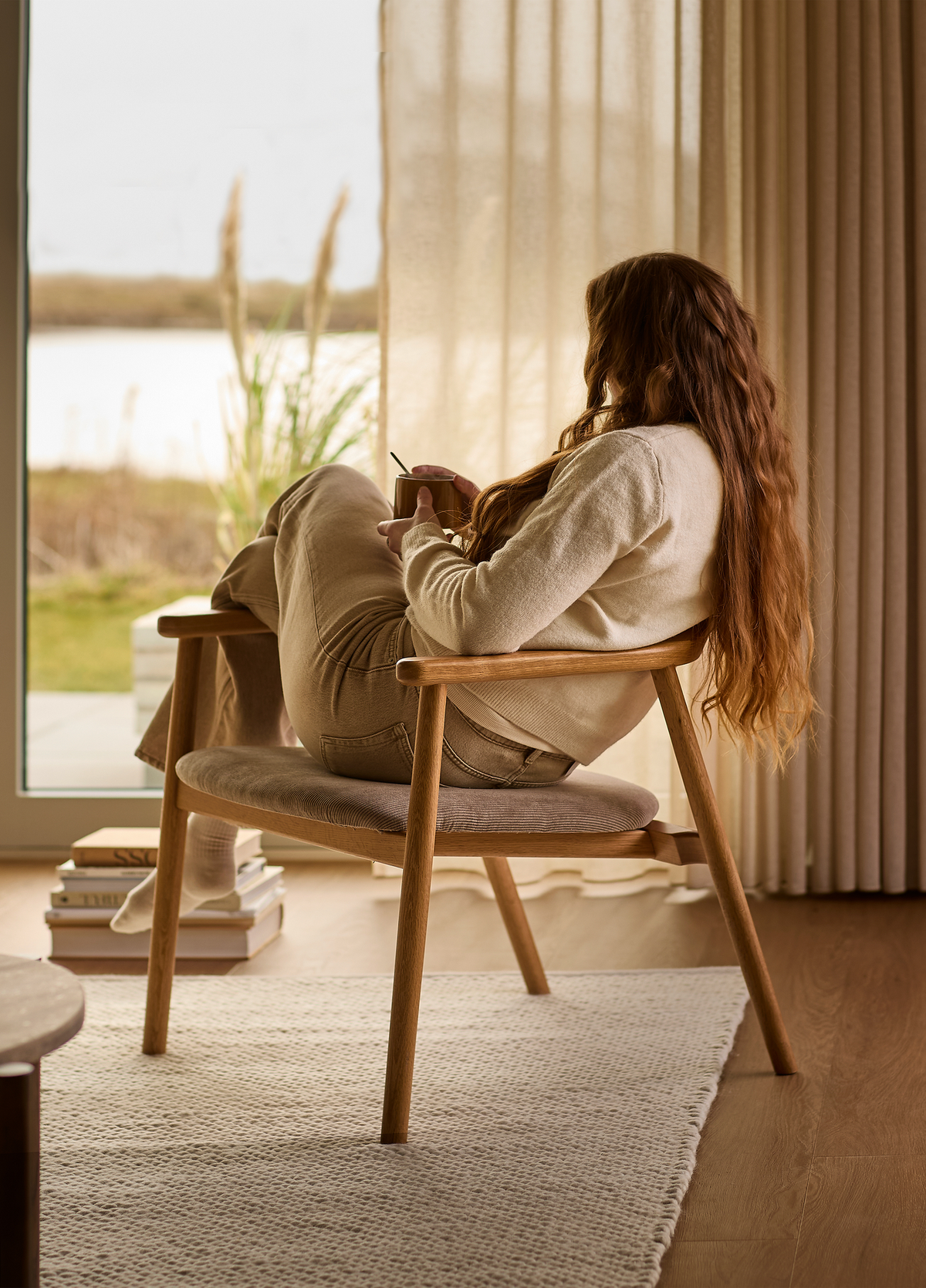 Person i loungestol ved vindue med kop, bøger på gulvet, lyse gardiner og neutral tæppe i en hyggelig stue.