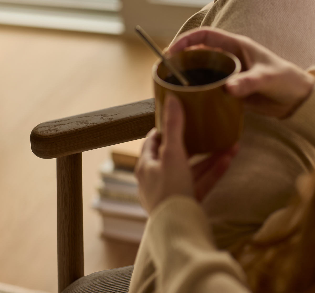 Nærbillede af hænder, der holder en brun kop med ske, siddende i en trælænestol ved siden af en stak bøger i et varmt, lyst stueområde.