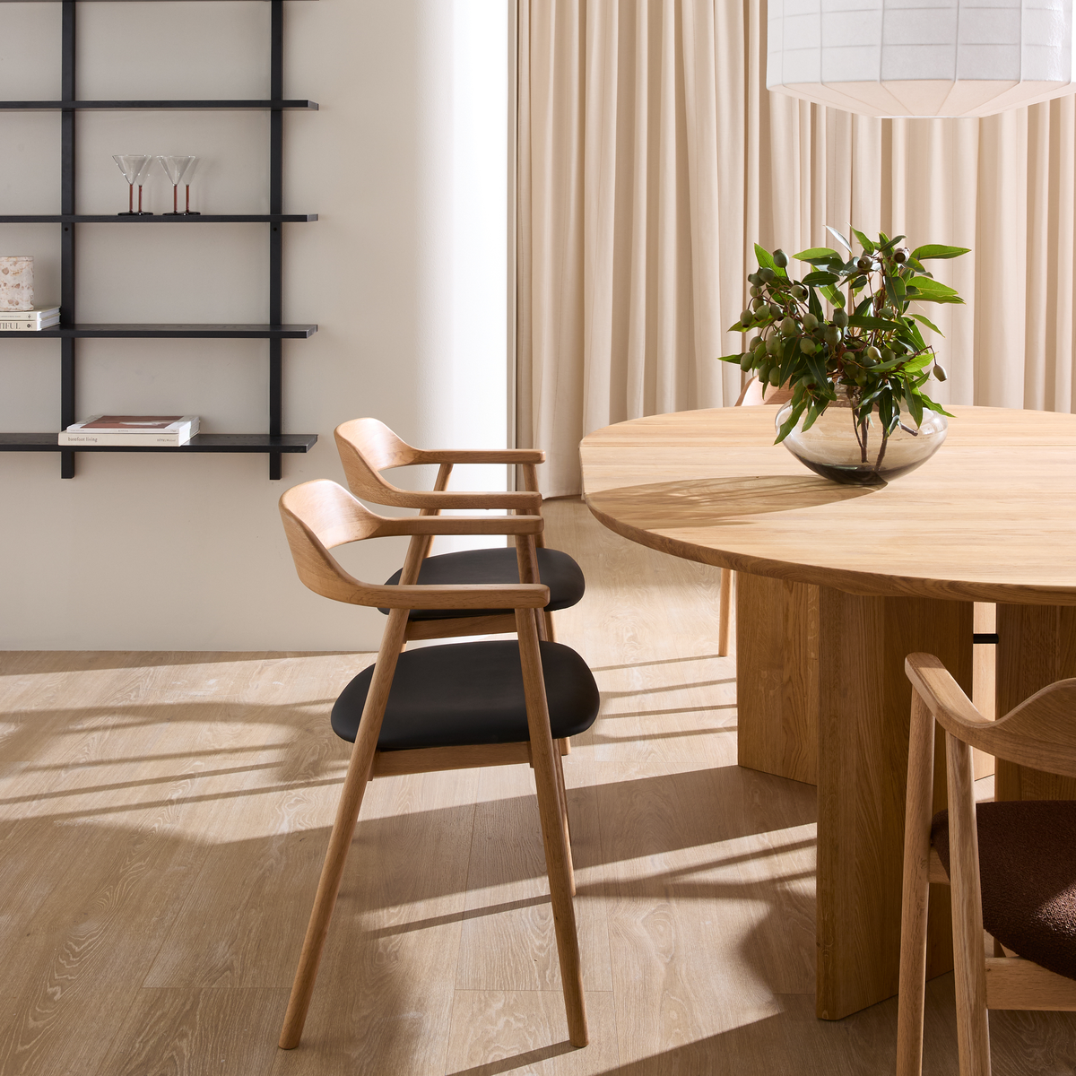 Lyst, minimalistisk spisestue med rundt egetræsbord, træstole med sorte sæder, sort væghylde med glas og bøger, beige gardiner, hvid pendellampe og glasvase med grønne grene.