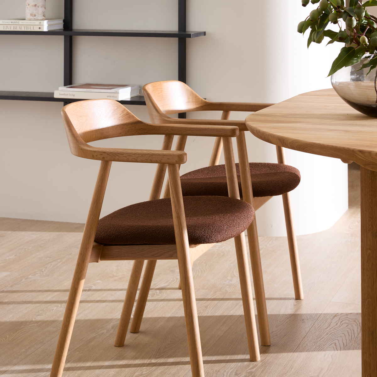 To lyse træstole med brune sæder ved et egetræsbord, bøgerne Live Beautiful og Barefoot living på væghylden og en grøn plante i glasvase i et lyst, minimalistisk spisemiljø.