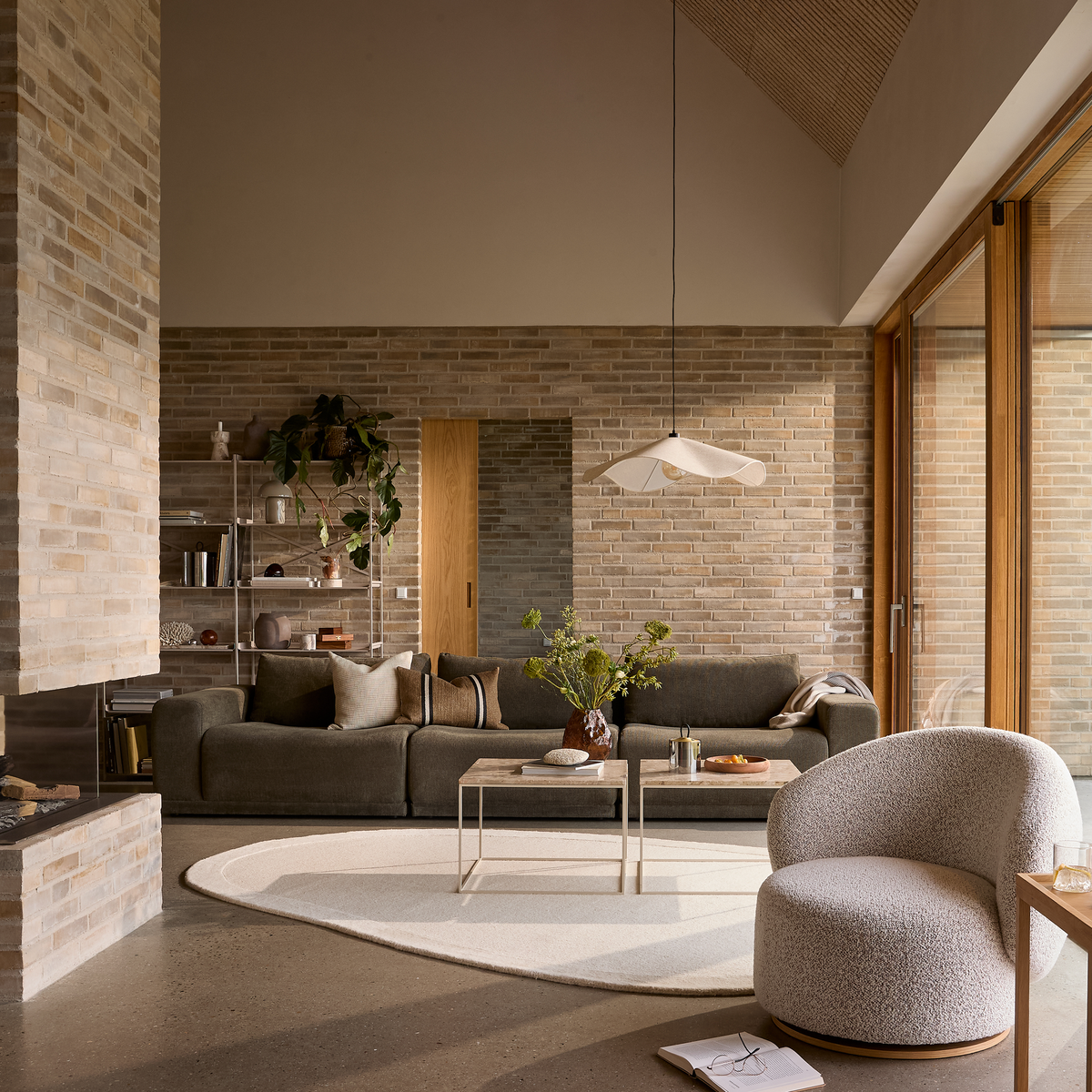 Stue med brun sofa, lyst ovalt tæppe og hvid loungestol; to sofaborde med vase med grønne grene under en skulpturel pendellampe foran murstensvæg og store vinduer.