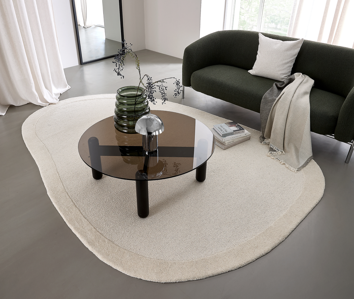 Moderne stue med mørkegrøn sofa, organisk cremefarvet tæppe og rundt sofabord i tonet glas, med grøn glasvase med gren og lille krombordlampe; spejl og lyse gardiner i baggrunden.
