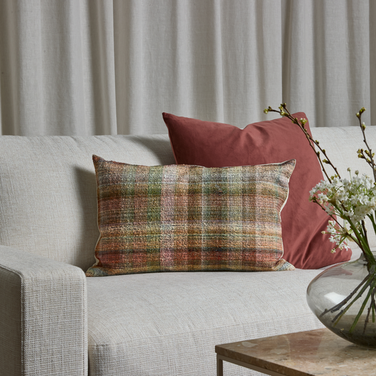 Beige sofa med en aflang pude i varme tern og en rustfarvet pude; glasvase med hvide blomster på stenbord, lyse gardiner i baggrunden.