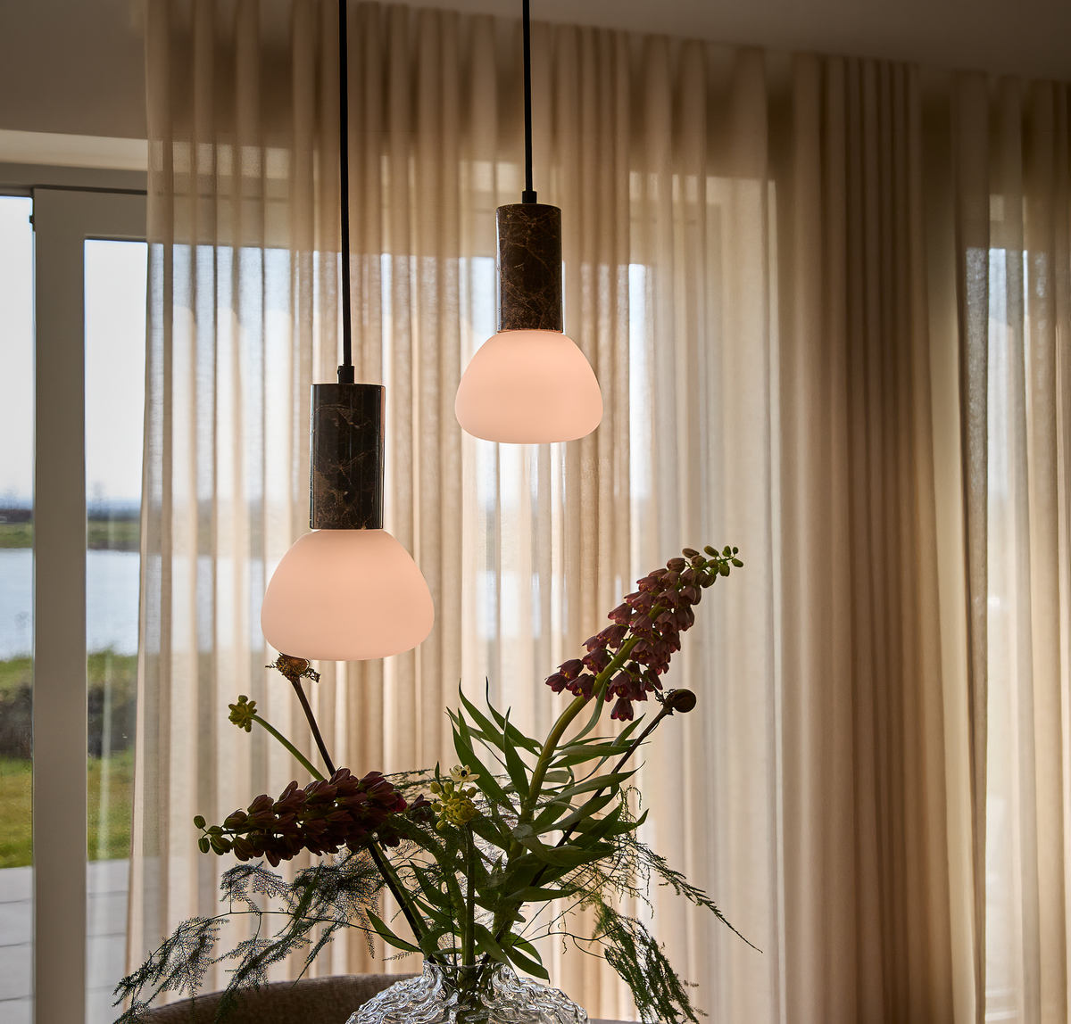 To pendler med opalglas over en vase med blomster foran beige gardiner og et vindue i dagslys.