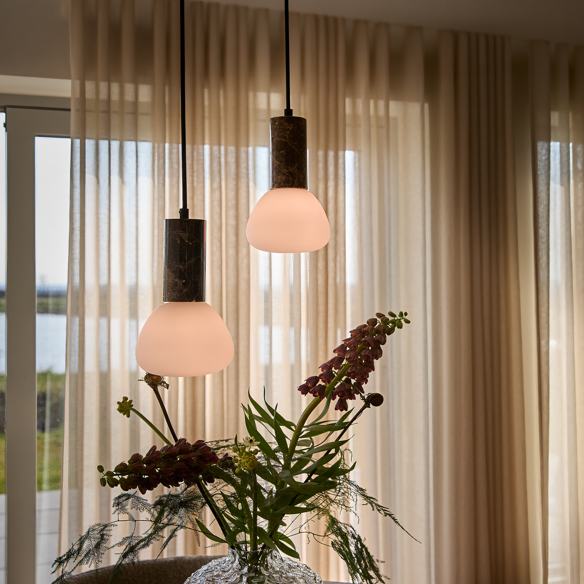 To moderne pendellamper med bløde opalglasskærme hænger foran beige, transparente gardiner over en buket i glasvase ved et vindue.