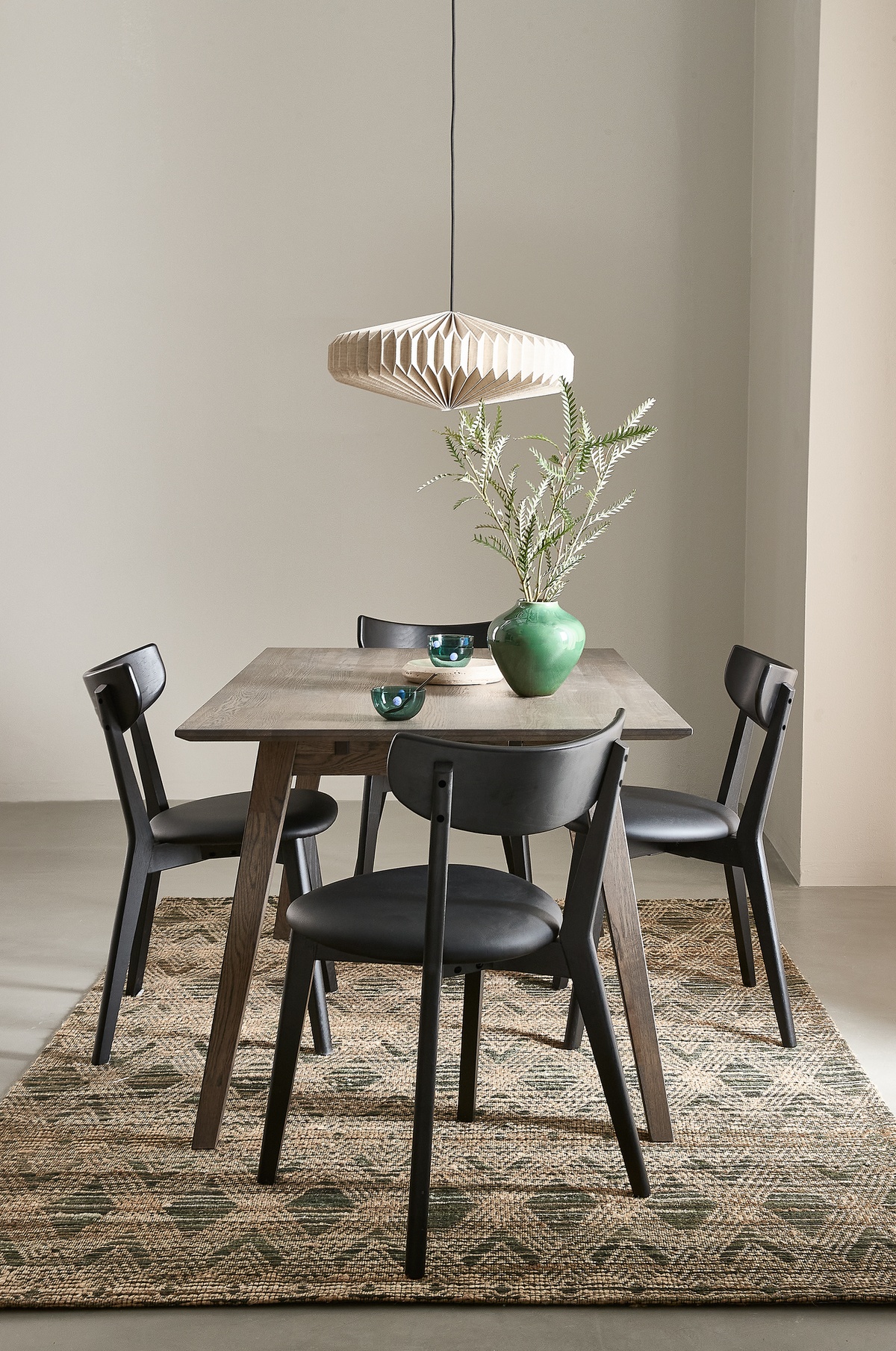 Spiseplads med træbord, fire sorte stole, foldet pendel og grøn vase med grene på et mønstret naturfarvet tæppe i en lys, minimalistisk indretning.