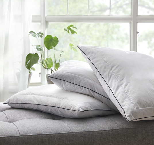 To hvide puder med sort piping stablet på en grå seng foran et lyst vindue med grønne planter.