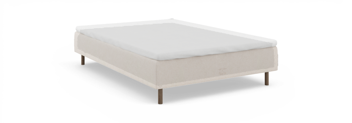 Seng med beige polstret ram, hvid topmadras og træben – set skråt forfra på sort baggrund.