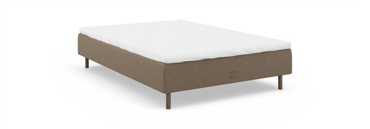 Sengebund i beige tekstil med hvid topmadras og runde træben, frilagt.