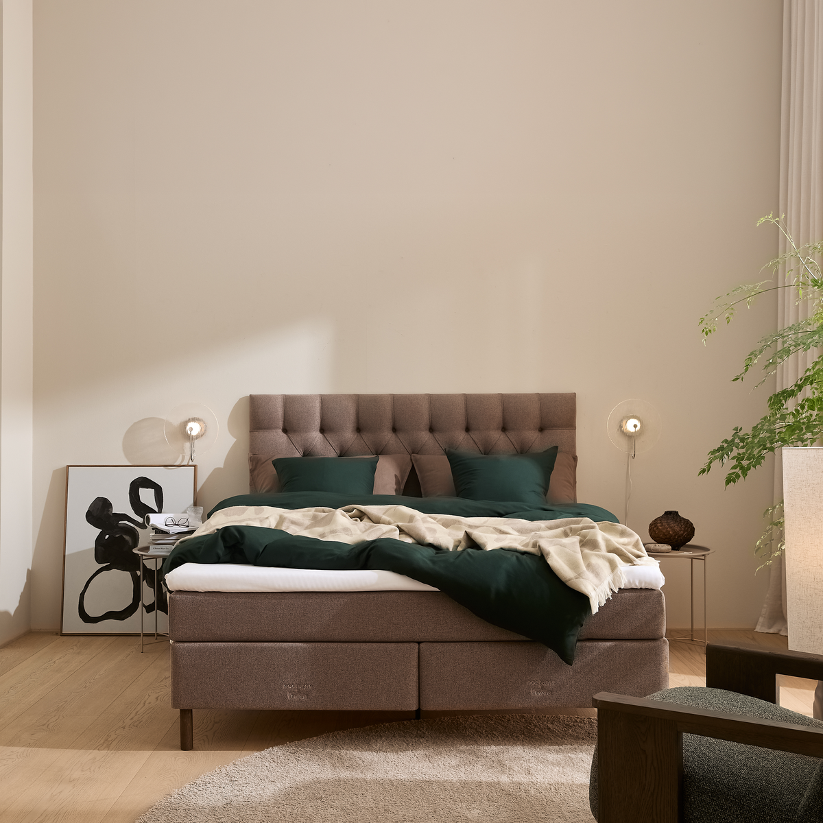 Stilrent soveværelse med polstret sengegavl, grønt sengesæt og beige plaid; væglamper, kunst print op ad væggen, lille sidebord med bøger og grøn plante i hjørnet.