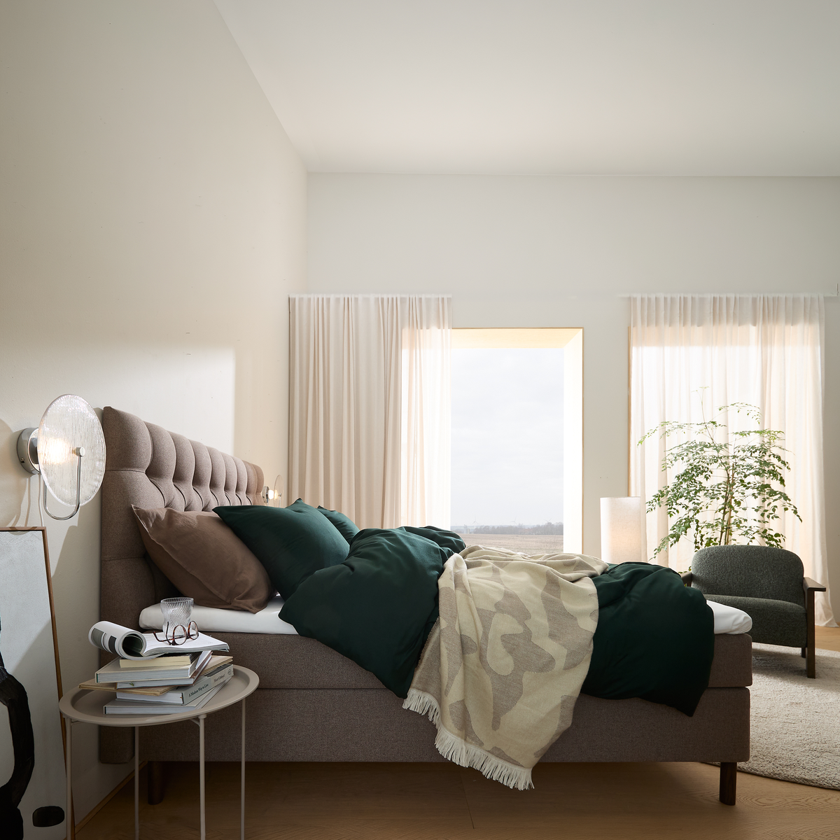 Lyst soveværelse med polstret seng, grønne puder og plaid, store lette gardiner, gulvlampe og plante ved vinduet.
