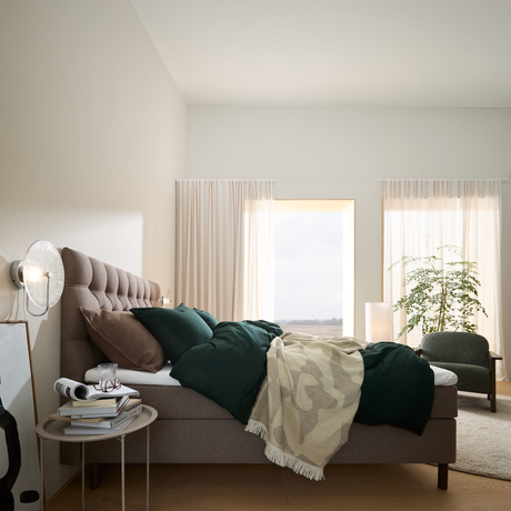Lyst soveværelse med polstret seng, grønne puder og plaid, store lette gardiner, gulvlampe og plante ved vinduet.