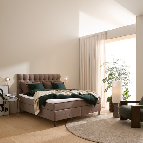 Lyst soveværelse med beige vægge, tuftet sengegavl, grønne puder og dyne, rund beige tæppe, lyse gardiner, væglamper, gulvlampe og stor grøn plante ved vinduet.
