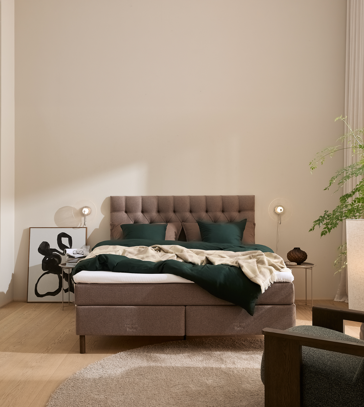 Seng med polstret gavl, mørkegrønt sengetøj og beige plaid i lyst skandinavisk soveværelse med væglamper, tæppe og grøn plante.
