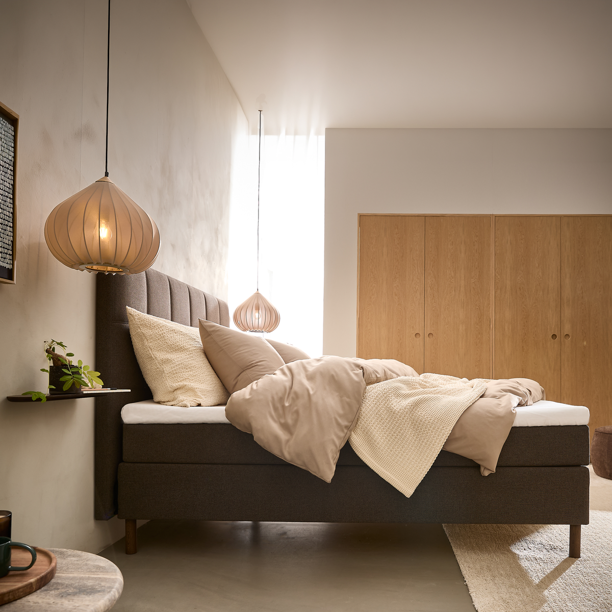Lyst soveværelse med polstret brun seng, beige sengetøj, to pendler og garderobeskabe i lyst træ.