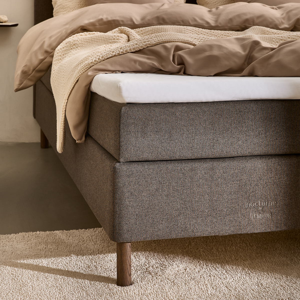 Grå stofbetrukket seng med hvid topmadras og beige dyner; logoet nocturne TEMPUR på sengekanten.