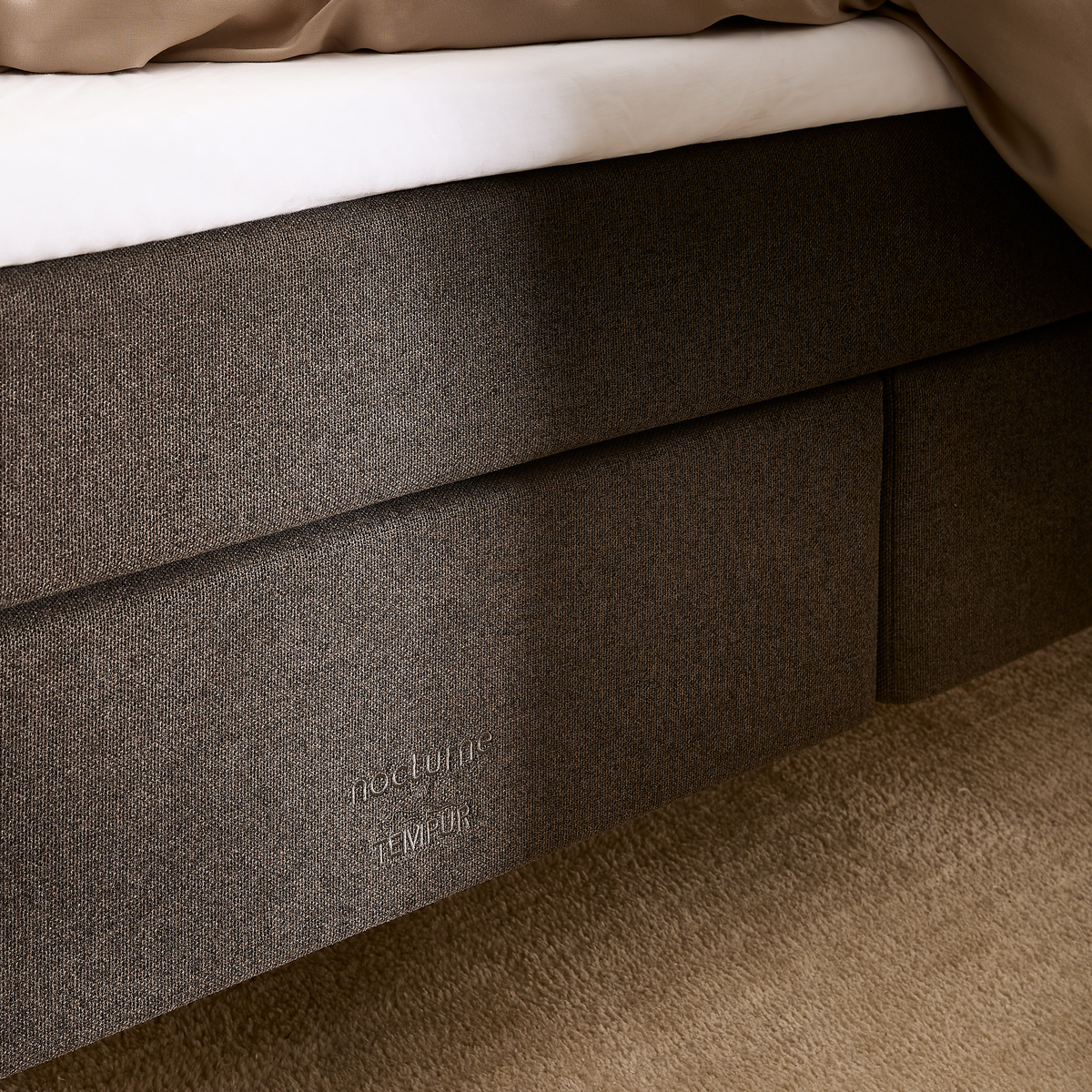 Nærbillede af en mørk stofpolstret sengbund med “nocturne TEMPUR”-branding, hvid topmadras og beige sengetæppe på beige tæppe i et soveværelse.
