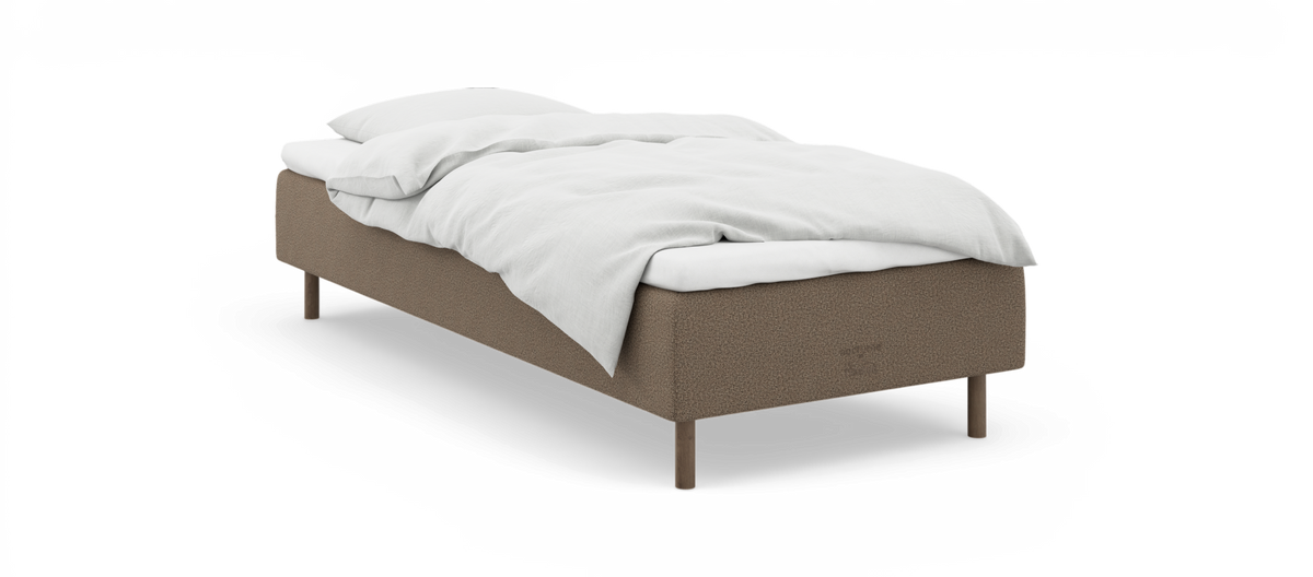 En enkeltseng med hvidt sengetøj på beige polstret sengeramme med træben, fotograferet skråt forfra mod hvid baggrund.