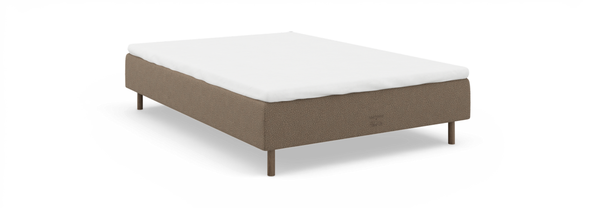 Enkel seng med hvid topmadras på beige stofsengebase med træben, set skråt forfra på sort baggrund.