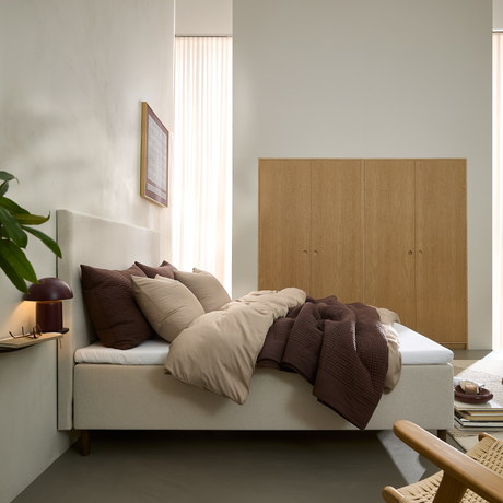 Soveværelse med beige seng, brune og beige dyner og puder, lyse træskabe og blødt dagslys gennem skærmede gardiner; bordlampe og plante ved siden af.